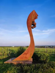 Eine moderne Skulptur in einem Weinberg unter einem klaren Himmel. Die Skulptur hat eine geschwungene Form und trägt runde Elemente.