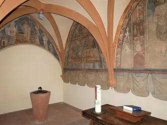 Ein historischer Raum mit kunstvollen Wandmalereien und einem Brunnen in der Mitte. Der Boden ist mit Fliesen bedeckt und es gibt einen Holztisch mit einem Buch.