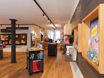 Ein gemütlicher Buchladen mit Holzregalen und warmem Licht. Bücher sind ordentlich auf Regalen und Tischen arrangiert.