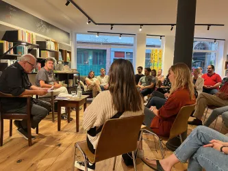 Eine Buchlesung in einem gemütlichen Raum mit Sitzgelegenheiten. Menschen hören gespannt zu und nehmen an der Veranstaltung teil.