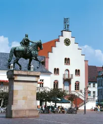 Ein Reiterstandbild auf einem Sockel steht im Vordergrund. Im Hintergrund ist ein historisches Gebäude mit einer auffälligen Uhr und einem klaren blauen Himmel.