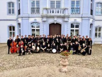 Eine große Musikgruppe steht vor einem schönen, historischen Gebäude. Die Mitglieder tragen schwarze Kleidung und halten verschiedene Musikinstrumente.