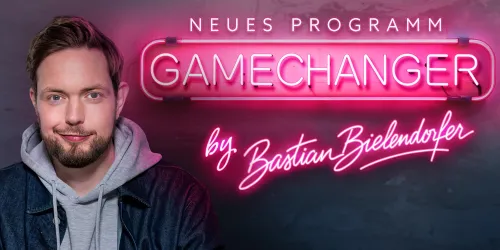 Ein Mann lächelt vor einem neonbeleuchteten Hintergrund mit dem Schriftzug "Gamechanger". Er trägt einen Hoodie und eine Jeansjacke.