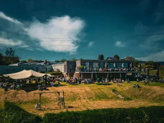 Eine lebhafte Veranstaltung im Freien mit vielen Menschen, die auf einer Terrasse stehen. Im Vordergrund sind eine Hängematte und ein Spielplatz sichtbar.