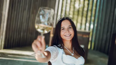 Eine junge Frau in traditioneller Tracht hält ein Glas Sekt und lächelt. Im Hintergrund sind Holzstrukturen und sanftes Licht zu sehen.