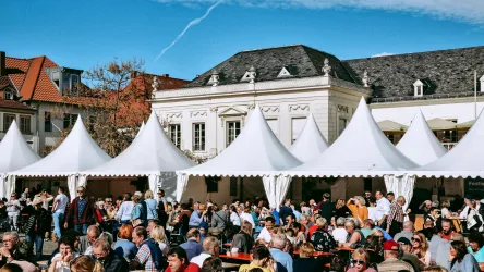Ein belebter Markt mit vielen Menschen, die an Tischen sitzen. Zelte mit weißen Dächern sind im Hintergrund zu sehen, und der Himmel ist klar und blau.