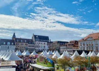 Ein lebhafter Marktplatz mit vielen Verkaufsständen und Bürgern. Der Himmel ist blau mit einigen Wolken.
