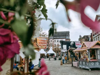 Ein festlicher Markt mit bunten Ständen und Zelten in einer Stadt. Umgeben von Blumen und einem wolkigen Himmel.