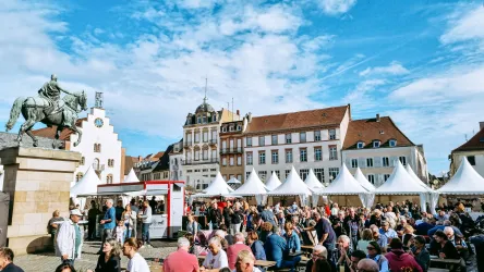 Ein belebter Platz mit vielen Menschen und festlichen Zelten. Im Hintergrund sind historische Gebäude und ein Reiterdenkmal zu sehen.
