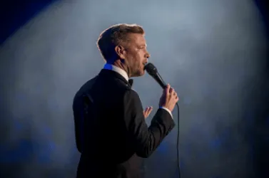 Ein Mann in einem Anzug steht auf der Bühne und spricht in ein Mikrofon. Im Hintergrund ist ein sanftes Licht zu sehen.