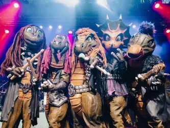 Fünf Dinosaurier-Maskottchen in bunten Kostümen stehen auf der Bühne. Sie halten Instrumente und scheinen Teil einer musikalischen Aufführung zu sein.