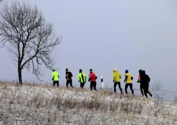 Eine Gruppe von Läufern joggt auf einem verschneiten Hügel. Im Hintergrund steht ein einzelner Baum unter einem grauen Himmel.