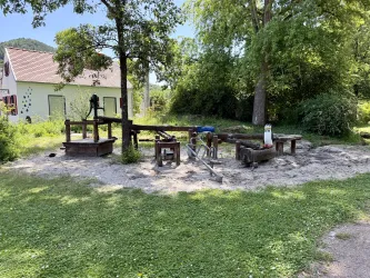 Ein Spielplatz mit wenigen Spielgeräten aus Holz steht in einem sandigen Bereich. Im Hintergrund sind Bäume und ein Gebäude zu sehen.