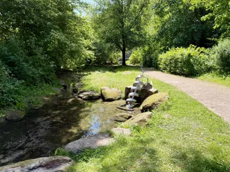 Ein idyllischer Weg entlang eines kleinen Bachs. Grüne Bäume und Sträucher umgeben den Bereich, während ein Wasserspiel aus Steinen im Vordergrund zu sehen ist.