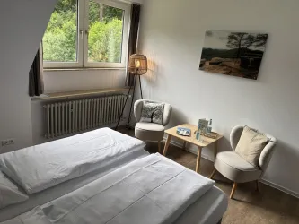 Ein helles Zimmer mit zwei Betten, einem kleinen Tisch und gemütlichen Sesseln. Große Fenster bieten einen Blick ins Grüne.