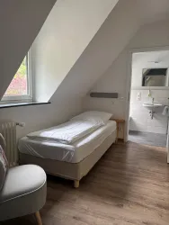 Ein einfaches Schlafzimmer mit einem Einzelbett und einem Stuhl. Durch das Fenster scheint Tageslicht und im Hintergrund ist ein Badezimmer sichtbar.