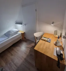 Ein einfaches Zimmer mit einem Bett und einem Schreibtisch. Es gibt eine Lampe und einige Wasserflaschen auf dem Tisch.