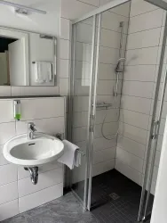 Ein modernes Badezimmer mit einer Dusche aus Glas und einem Waschbecken. Die Wände sind hell gefliest und es gibt einen Spiegel über dem Waschbecken.