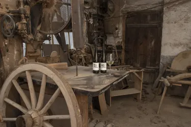 Ein alter Raum mit einer rustikalen Holzwerkbank und verschiedenen Werkzeugen. Auf der Tischplatte stehen drei Flaschen mit Weinetiketten.