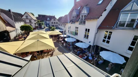 Eine lebhafte Außenterrasse mit vielen Gästen unter Sonnenschirmen. Umgeben von charmanten Gebäuden in einer sonnigen Umgebung.