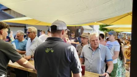 Eine belebte Freiluftveranstaltung mit vielen Menschen, die an einem Thekenbereich stehen. Sonnenschirme schützen vor der Sonne, während die Gäste miteinander sprechen und Getränke genießen.