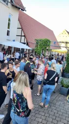 Eine lebhafte Menschenmenge auf einer gepflasterten Straße, umgeben von Gebäuden und Sonnenschirmen. Viele Gäste genießen das Wetter und unterhalten sich.