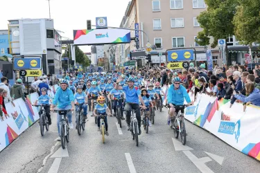 Eine große Gruppe von Radfahrern in blauen T-Shirts fährt auf einer Straße. Viele Zuschauer stehen an den Seiten und feuern sie an.