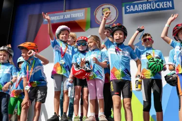 Eine Gruppe von Kindern steht auf einer Bühne und feiert ihre Erfolge. Sie tragen Helme und T-Shirts mit bunten Motiven und haben Medaillen um den Hals.