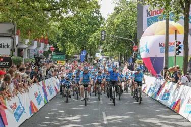 Eine Gruppe von Radfahrern nimmt an einem Event teil und fährt durch eine Straße, während Zuschauer am Straßenrand zuschauen. Bunte Werbebanner und ein großer, aufblasbarer Ball sind im Hintergrund zu sehen.