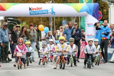 Kinder fahren mit ihren Fahrrädern unter einem bunten Zielbogen. Zuschauer jubeln und feiern an der Strecke.