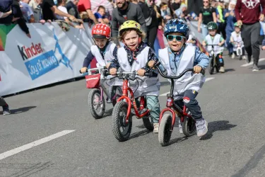 Eine Gruppe von Kindern fährt mit kleinen Fahrrädern in einem Wettkampf. Die Begeisterung der Zuschauer im Hintergrund ist deutlich zu spüren.