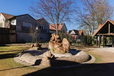 Ein schöner Brunnen in einem Garten mit Wohnhäusern im Hintergrund. Die Umgebung ist grün, und Bäume sind sichtbar.