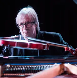 Ein Musiker sitzt am Klavier und spielt eine Violine. Der Fokus liegt auf seiner konzentrierten Miene und der Instrumente.