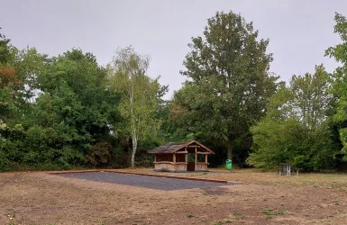 Eine kleine, rustikale Hütte steht in einem offenen Bereich, umgeben von Bäumen. Der Boden ist trocken und unauffällig, mit einem gepflasterten Bereich in der Mitte.