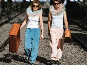 Ein Paar in auffälligen Outfits mit großen Kragen geht auf Gleisen. Jeder trägt einen Koffer in der Hand.