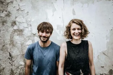 Ein lächelndes Paar steht vor einer grauen Wand. Beide tragen lässige Kleidung und wirken freundlich und entspannt.