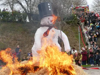 Ein großer Schneemann mit einem Zylinder wird verbrannt. Im Hintergrund stehen viele Zuschauer, die das Ereignis beobachten.