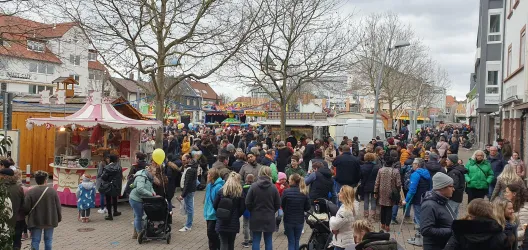 Ein belebter Marktplatz mit vielen Menschen und Ständen. Es gibt fröhliche Atmosphäre und verschiedene Verkaufsstände.