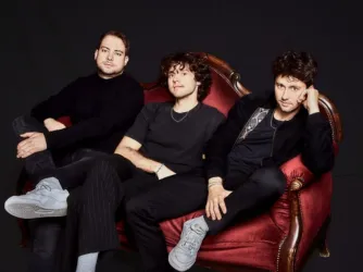 Drei Musiker sitzen entspannt auf einer roten Couch. Sie tragen schwarz-weiße Kleidung und wirken stilvoll und modern.