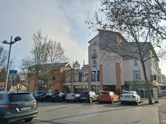 Ein modernes Gebäude mit mehreren Stockwerken und großen Fenstern. Geparkte Autos stehen auf einem Platz vor dem Gebäude, umgeben von einigen Bäumen.