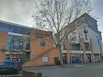 Ein modernes Gebäude mit großen Fenstern und einer zentralen Treppe. Ein Baum steht vor dem Gebäude, das eine einladende Atmosphäre schafft.