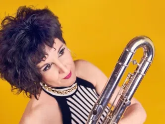Eine Frau mit lockigem Haar hält ein großes Blasinstrument in der Hand. Der Hintergrund ist leuchtend gelb, was eine fröhliche Atmosphäre schafft.