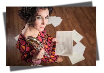 Eine Frau hält ein Musikinstrument in der Hand und schaut direkt in die Kamera. Um sie herum liegen Notenblätter auf dem Boden.