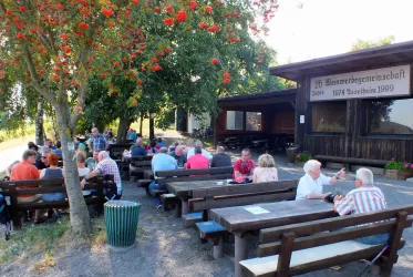 Eine gesellige Gruppe von Menschen sitzt an Holztischen im Freien. Im Hintergrund ist ein Gebäude mit einem Schild zu sehen.