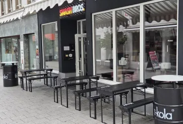 Eine Straßenszene mit mehreren schwarzen Tischen und Stühlen vor einem Restaurant. Das Fenster des Restaurants ist groß und einladend.