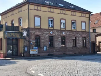 Ein historisches Gebäude mit einem gelben Fassadenanstrich und großen Fenstern. Es befindet sich an einer Straßenkurve und hat eine steinerne Umgebung.
