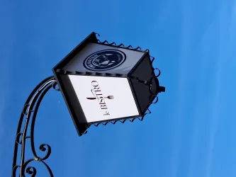 Ein hängendes Schild mit der Aufschrift "k Bistro" vor einem klaren blauen Himmel. Das Schild hat ein elegantes Design mit geschwungenen Details.