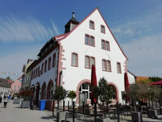 Ein charmantes, historisches Gebäude mit einer weißen Fassade und roten Details. Im Vordergrund sind Tische und Stühle auf einer Terrasse angeordnet.