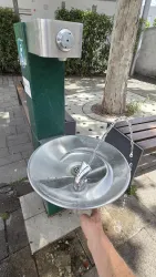 Ein Trinkbrunnen aus Metall steht auf einem Bürgersteig. Wasser sprudelt aus dem Hahn in die runde Schale.