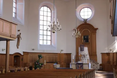 Eine helle Kirche mit hohen Fenstern und Holzinterieur. Der Altar ist im Vordergrund sichtbar, umgeben von Bänken.
